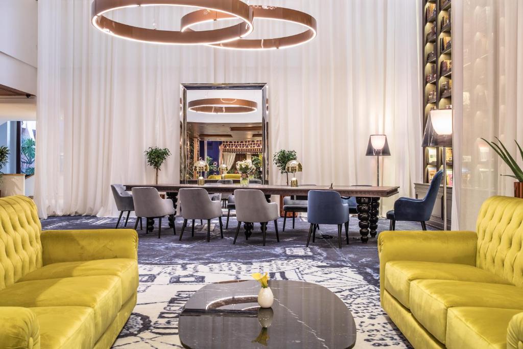 Mūsdienīgs viesnīcas interjers ar dzeltenām krēslām, elegantām lampām un lielu galdu, kas apņemts ar augu dekorācijām. Gaismas un tekstila kombinācija rada siltu un viesmīlīgu atmosfēru.