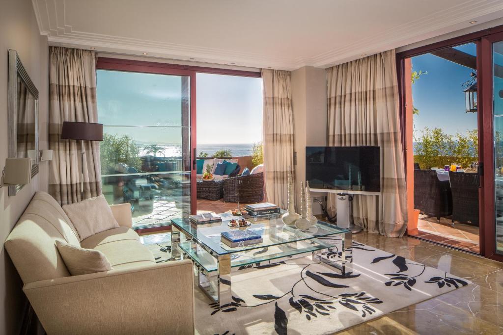 Mūsdienīga viesistaba ar skatu uz jūru, kurā ir eleganta dīvāna komplekts, stikla galdiņš un televizors. Lielie logi ļauj ieplūst dabiskajai gaismai un nodrošina piekļuvi terasei.