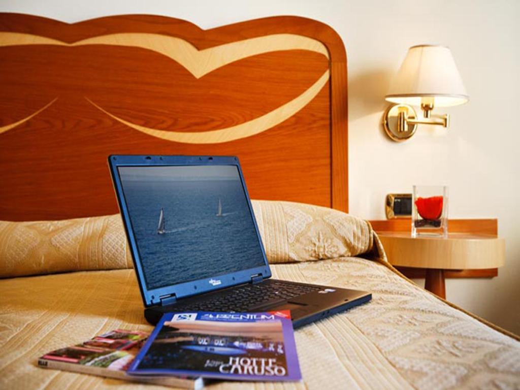 Klusa viesnīcas numura aina ar atvērtu klēpjdatoru, kas rāda burāšanas skatu, un žurnāliem uz gultas. Fons ar koka gultas galvgali un naktslampu.