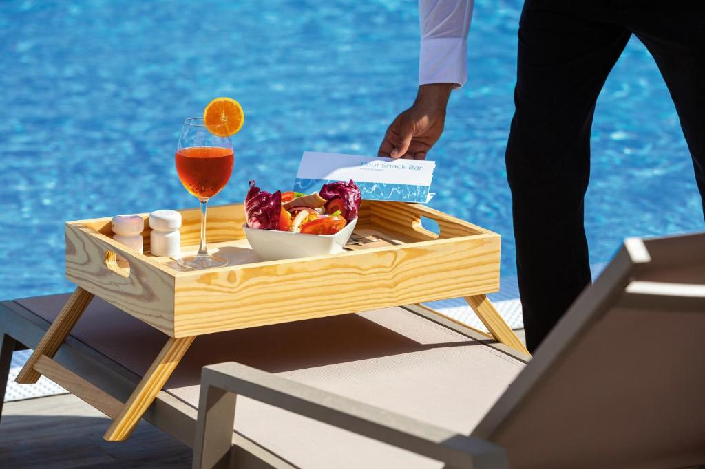 Koka paplāte ar dzērienu un uzkodu, kas tiek pasniegta pie baseina, kamēr redzams apkalpojošs personāls. Ideāla aina relaksācijai un brīvdienām.