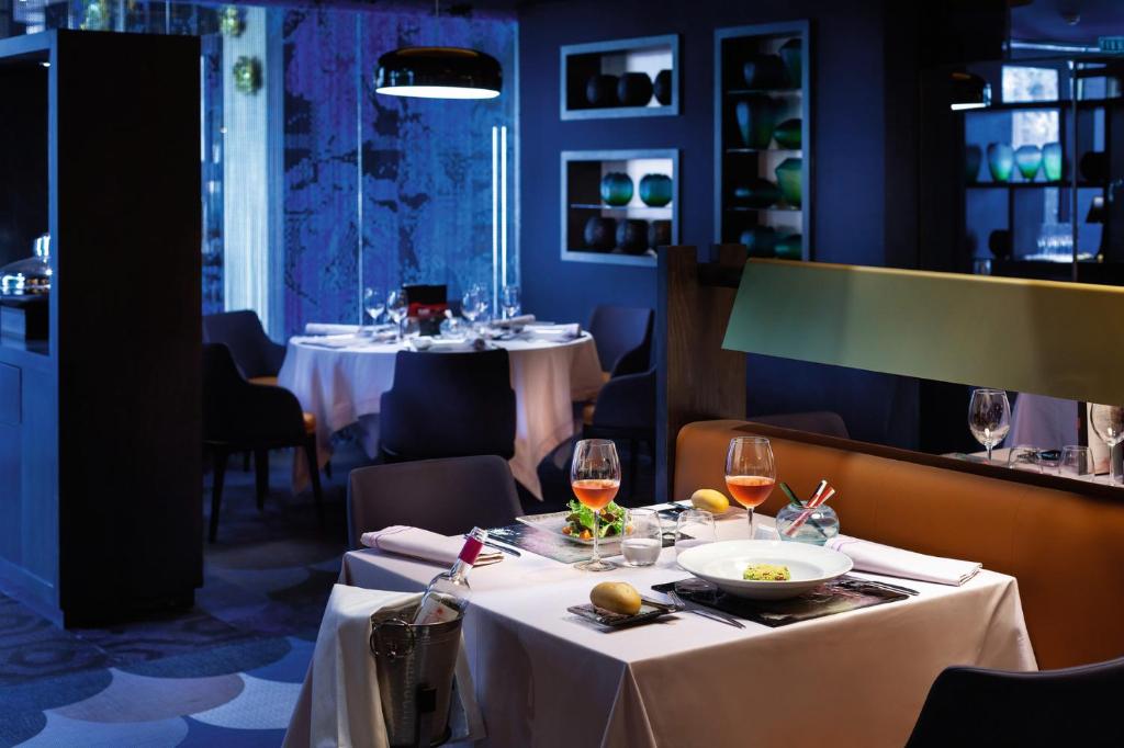 Elegants restorāns ar modernu interjeru, kurā redzami balti galdauti un glīti sakārtotas galda piederumi. Uz galda izvietota maltīte ar salātiem un dzērieniem, radot patīkamu atmosfēru vakariņām.