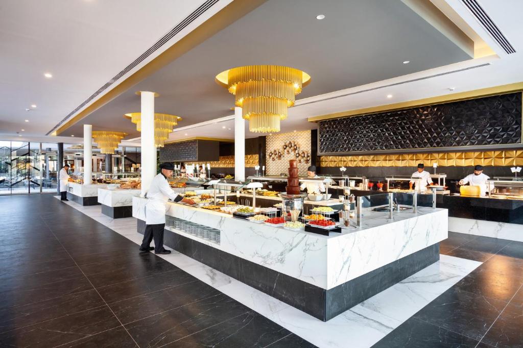 Mūsdienīgs restorāna interjers ar elegantu bufeti, kurā redzami šefpavāri, kas gatavo ēdienus. Telpā ir stilīgi gaismas elementi un moderni dekori.