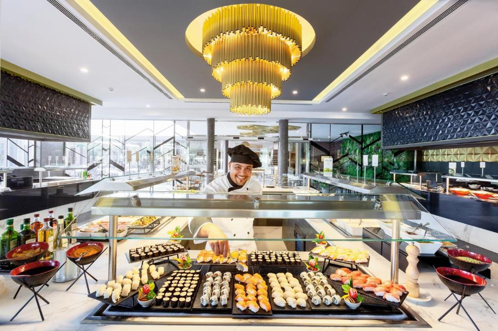 Sushi bārs ar šefpavāru, kas piedāvā dažādu suši veidu plāksnes, elegantā restorāna vidē ar modernu apgaismojumu un dizainu.