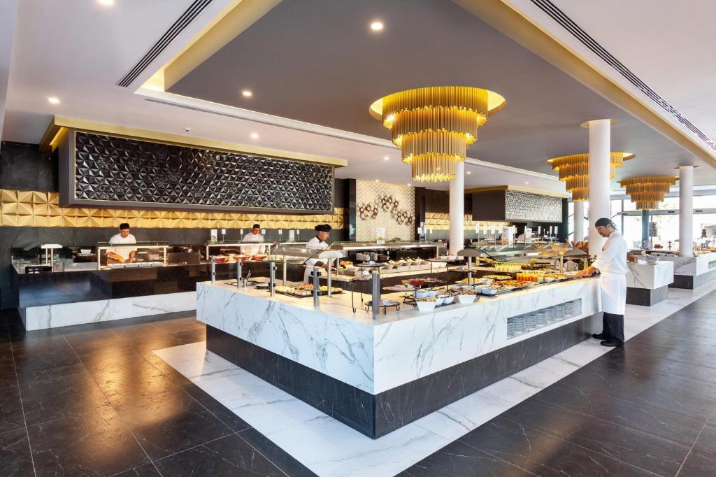 Moderns restorāns ar elegantu interjeru un bufeti, kur šefpavāri gatavo un pasniedz dažādus ēdienus. Gaismas elementi piešķir telpai siltumu un stilīgu izskatu.