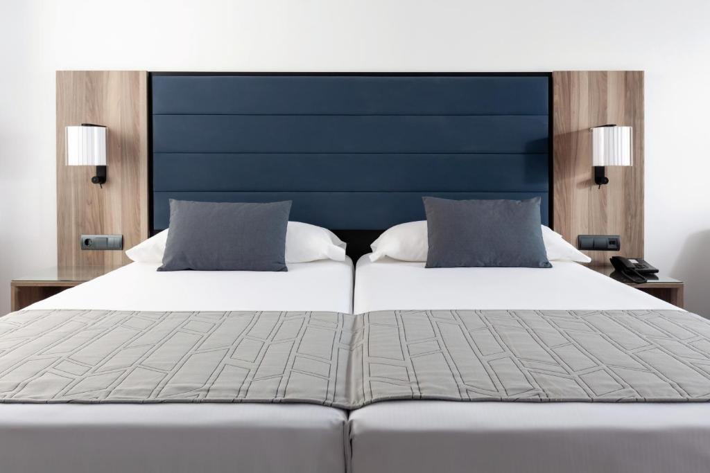 Mūsdienīgs guļamistabas interjers ar divvietīgu gultu, tumšzilu galvgali, pelēkiem spilveniem un koka naktsgaldiņiem. Gaismas pie sienas piešķir siltumu un komfortu.