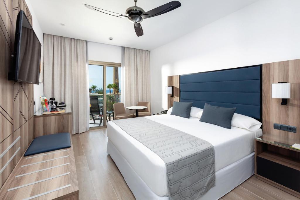 Moderna viesnīcas numura interjers ar gultu, TV un balkonu, no kura paveras skats uz dārzu. Numurs ir gaišs un elegants, ar koka akcentiem un neitrālām krāsām.