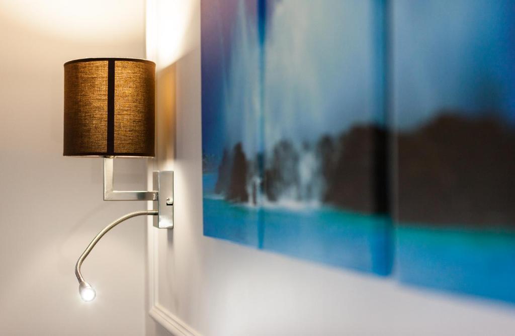 Sienas lampa ar brūnu abatmentu un sudraba stiprinājumu, blakus krāsainai ainavas glezniecībai. Interjera dizains rada modernu un elegantu atmosfēru.