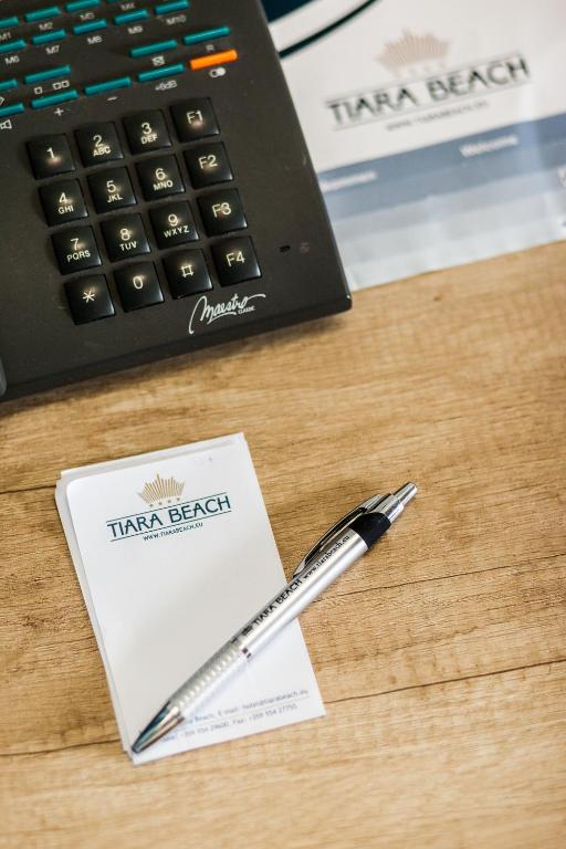 Tālruņa klaviatūra ar rakstāmbloku un pildspalvu uz koka virsmas, ar "Tiara Beach" logotipu uz papīra.