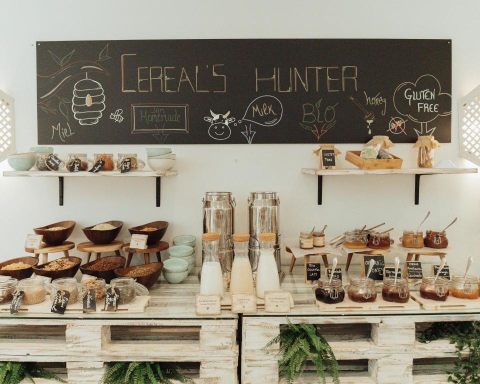 Svaigu brokastu stends ar dažādiem graudaugiem, pienu, medu un džemu, kas pasniegti koka traukos un stikla burkās. Uz sānu sienas redzama melna tāfele ar uzrakstu "Cereal's Hunter" un informāciju par pieejamajiem produktiem.