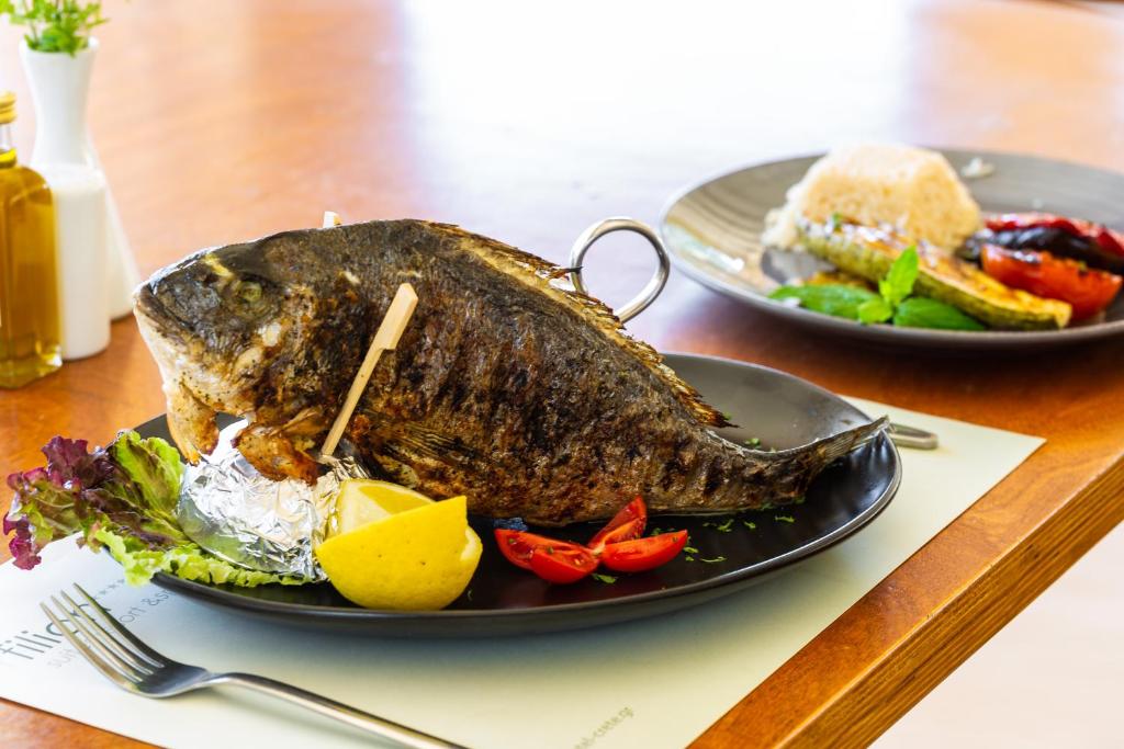 Grilēta zivs pasniegta uz melnas plates, kopā ar citronu, tomātiem un zaļumiem. Fons ietver citu ēdienu, tostarp rīsus un dārzeņus.