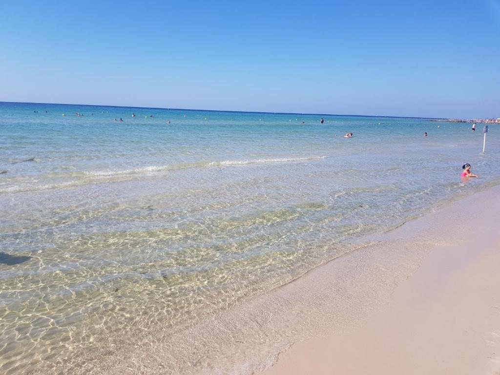Skats uz saulainu pludmali ar skaidru ūdeni un cilvēkiem, kas atpūšas un peldas. Siltā debesis un mierīga jūra rada relaksējošu atmosfēru.