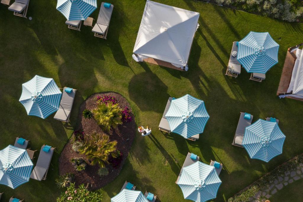 Lidojuma skats uz viesnīcas peldbaseina zonu ar pludmales gultām un strīpām, kas sniedz patvērumu. Apzaļumota teritorija ar ziediem rada relaksējošu atmosfēru.