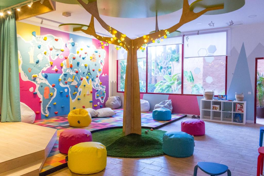 Krāsaina bērnu spēļu istaba ar kāpšanas sienu un mīkstajiem sēdekļiem. Centrā ir koks ar gaismiņām, radot draudzīgu atmosfēru.