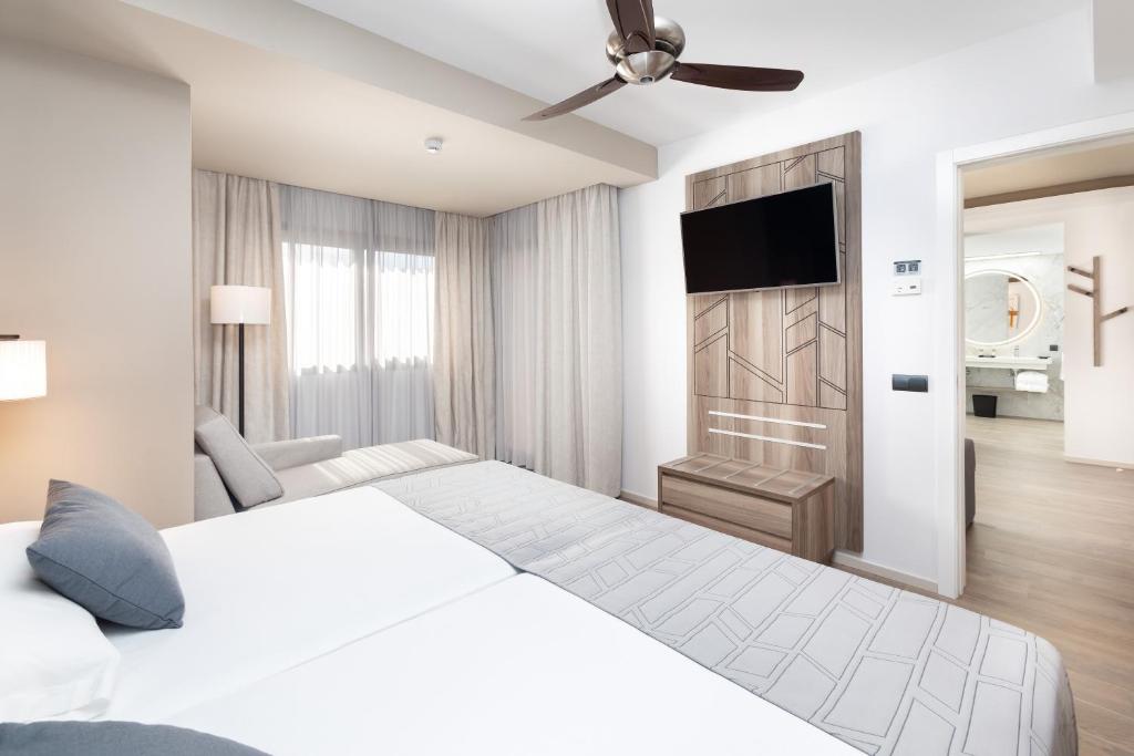 Mūsdienīgs viesnīcas numurs ar divām gultām, dīvānu un televizoru uz koka sienas, gaišas krāsas interjers ar dabīgu apgaismojumu.