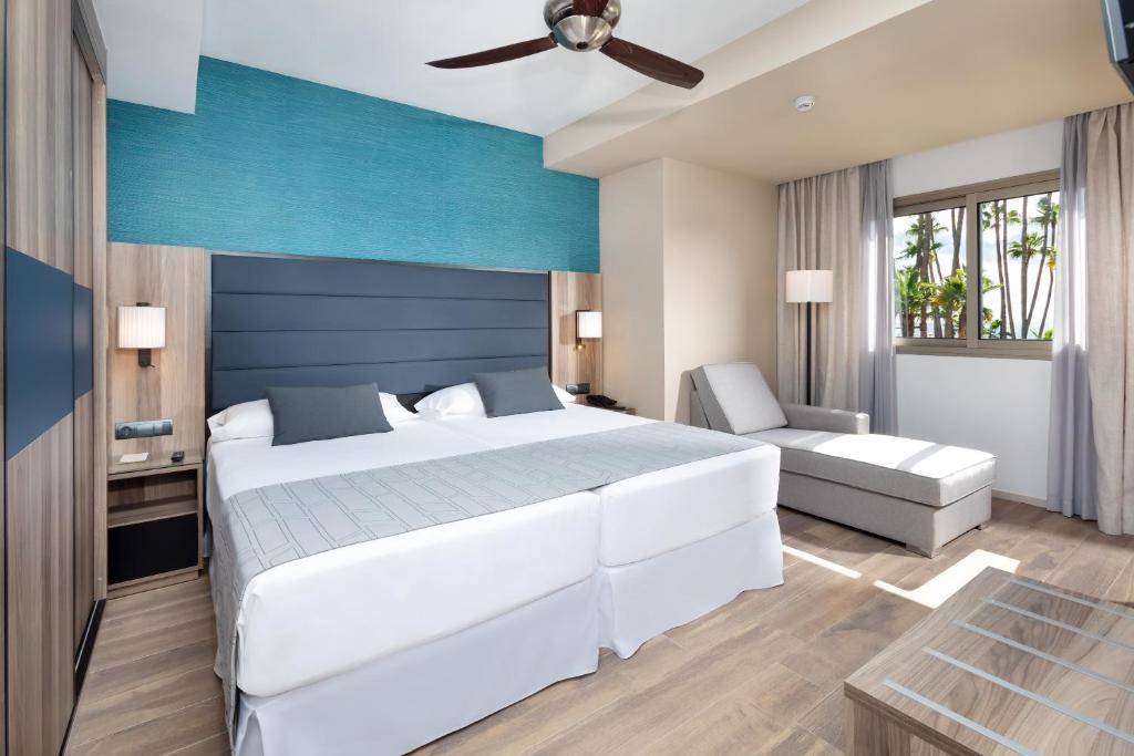 Moderns viesnīcas numurs ar divguļamo gultu, pelēkām gultas veļām un ērtu atpūtas krēslu. Interjers ir gaišs ar zilu akcentu un logu, caur kuru redzams ainava.