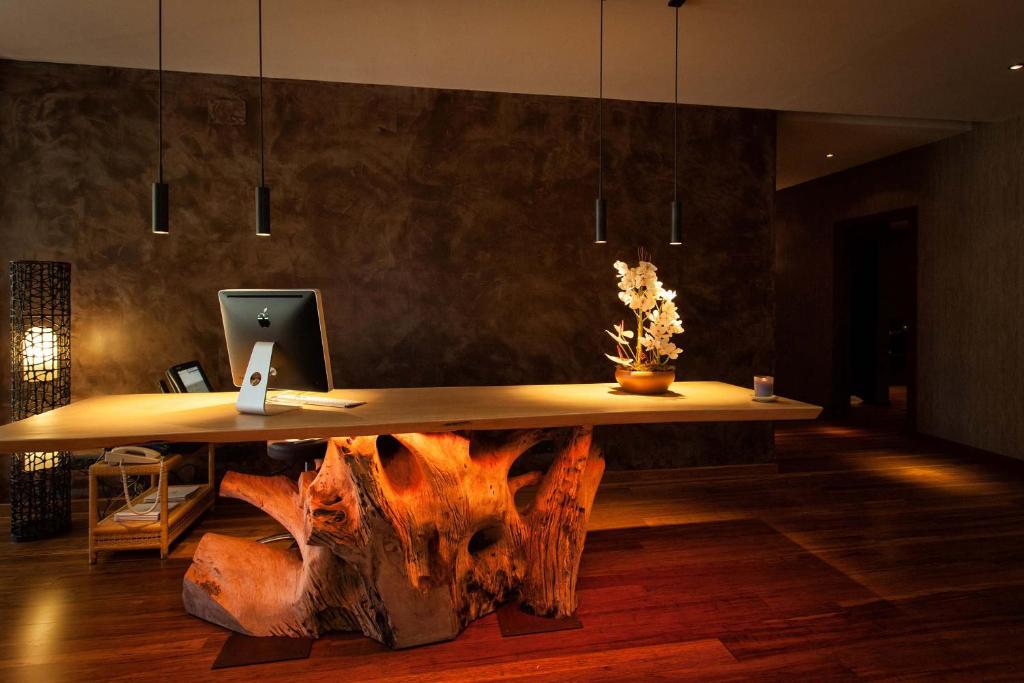 Mūsdienīgs biroja interjers ar koka galdu, uz kura novietota ziedu kompozīcija, un elegantu apgaismojumu. Pamatne ir izveidota no dabīga koka, radot siltu un aicinošu atmosfēru.