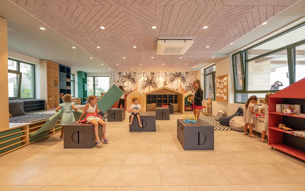 Bērnu rotaļu telpa ar moderniem elementiem, kur bērni spēlējas uz dažādām konstrukcijām un rotaļlietām. Telpā ir gaiša apgaismojuma, lieli logi un radošs interjers.