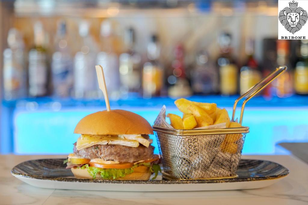 Gards burgers ar gaļas kotleti, sieru un dārzeņiem, pasniegts ar frī kartupeļiem. Fons ar dzērienu plauktu piešķir atmosfēru.