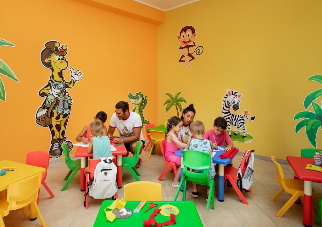 Krāsaina bērnu spēļu istaba ar jautriem zīmējumiem uz sienām, kur bērni un pieaugušie kopā nodarbojas ar radošām aktivitātēm pie krāsainiem galdiem.