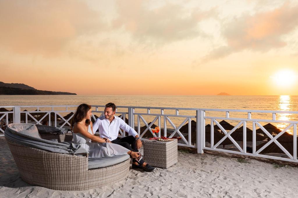 Romantiska pludmales ainava ar pāri, kas baudī dzīvespriecīgu brīdi saulrieta gaismā. Apsēdušies uz komfortabla dīvāna, viņi dalās brīnišķīgā atmosfērā ar kokteiļiem blakus.