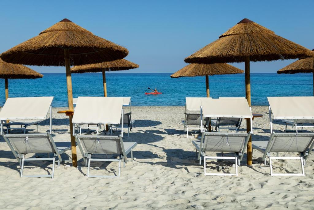 Pludmales ainava ar sauļošanās krēsliem un salmu jumtiem, kas izskatās uz mierīgu jūru, kurā redzami kajaku braucēji.