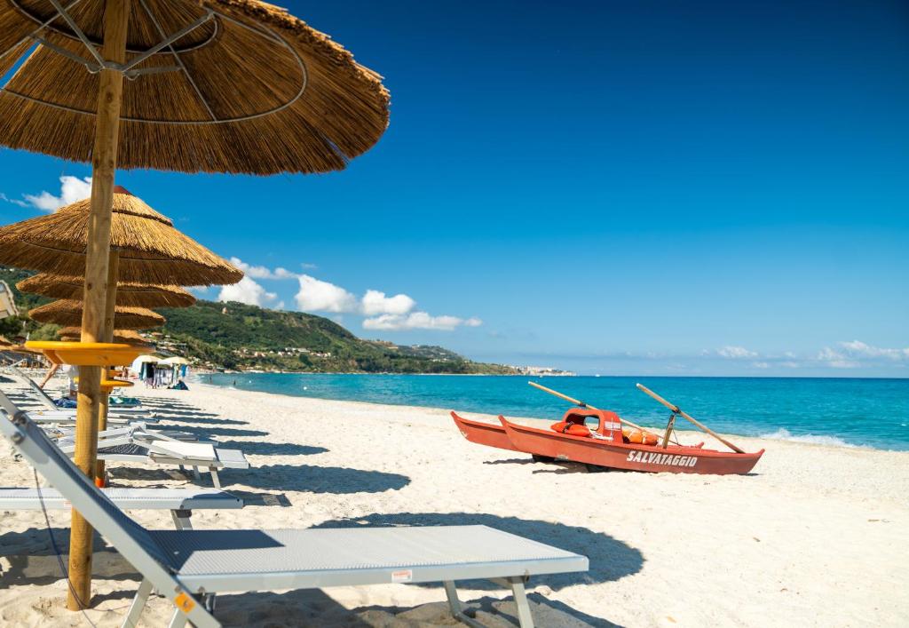 Pludmales ainava ar saulessargiem un sarkanu laivu, kas piestājusi pie smilšainā krasta, zem skaidra, zila debesīm.