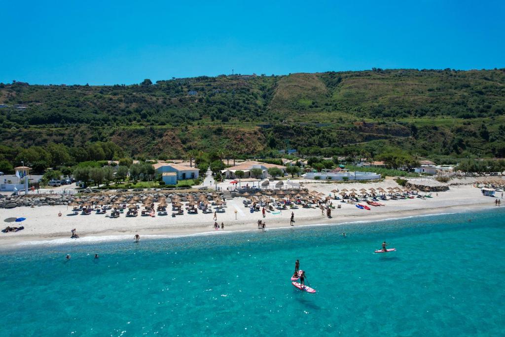 Kopējais skats uz pludmali ar saulessargiem un cilvēkiem, kas atpūšas pie ūdens, kā arī sērfošanas dēļiem jūrā. Zilā debesis un zaļās kalnu nogāzes veido idillisku ainavu.