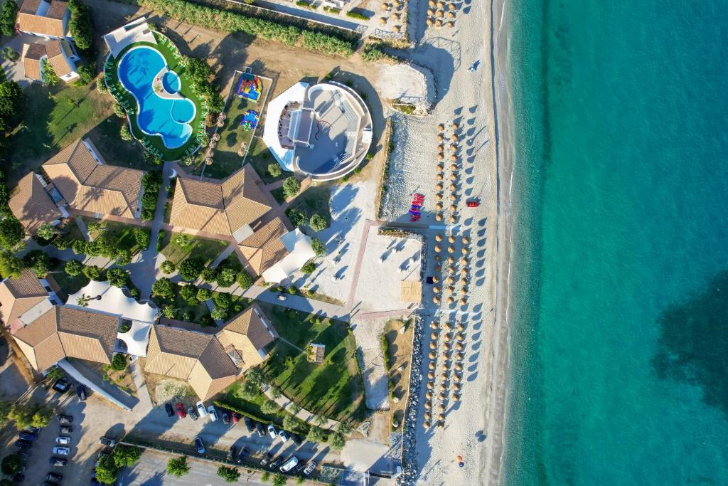 Gaisa skats uz kūrortu pie pludmales, ar baseinu, zaļām zonām un saulessargiem. Skaidrs zils ūdens un smilšu pludmale papildina mierīgu atmosfēru.