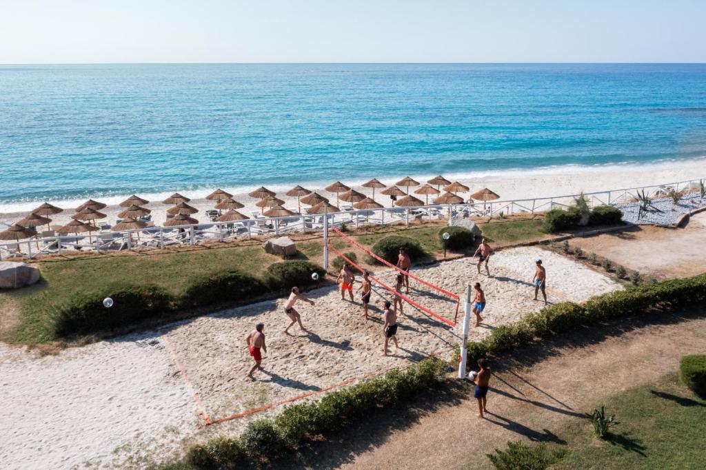 Pludmales volejbola spēle, kas notiek saulainā dienā pie jūras, ar sauļošanos un pludmales krēslām fonā.