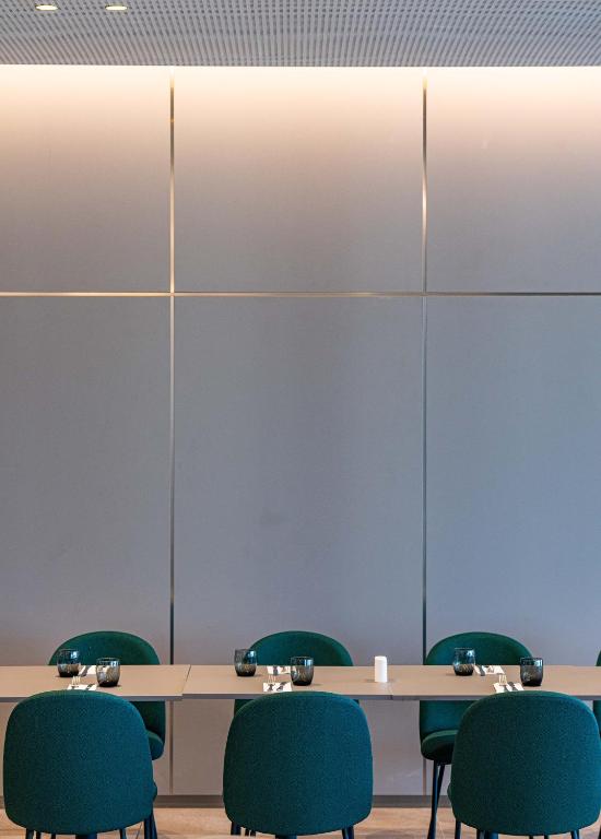 Mūsdienīga restorāna interjers ar zaļām krēsliem un elegantu galdu, kas izveidots pie sienas ar pelēku apdari. Ideāli piemērots relaksējošām vakariņām vai sanāksmēm.