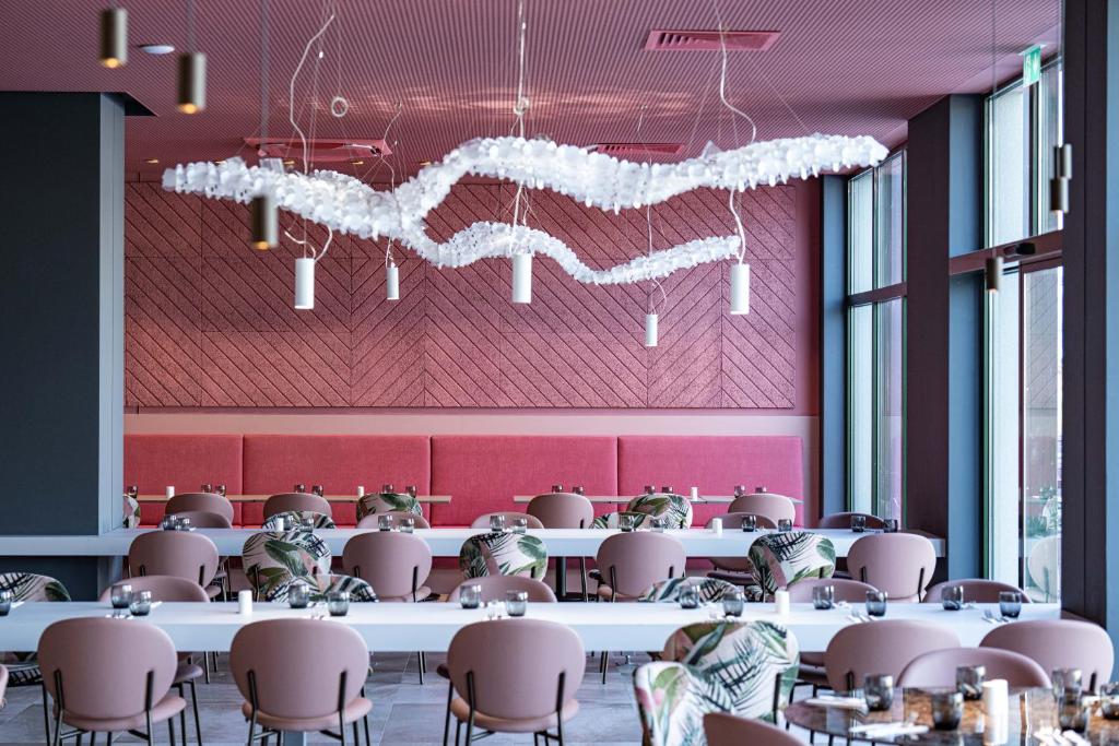 Mūsdienīga restorāna interjers ar rozā toņu sēdvietām un dekoratīvu gaismekli, kas rada patīkamu atmosfēru. Galdi ir sakārtoti ar elegantām detaļām un zaļiem akcentiem.