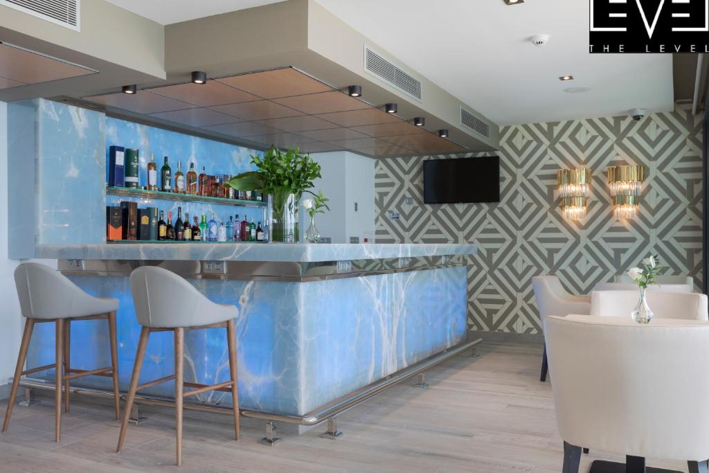 Mūsdienīgs bārs ar elegantu zilu marmora leti, aiz kuras redzami dažādi alkoholiskie dzērieni un ziedi. Interjerā ir moderns dizains ar ģeometriskām sienām un komfortablām krēsliem.