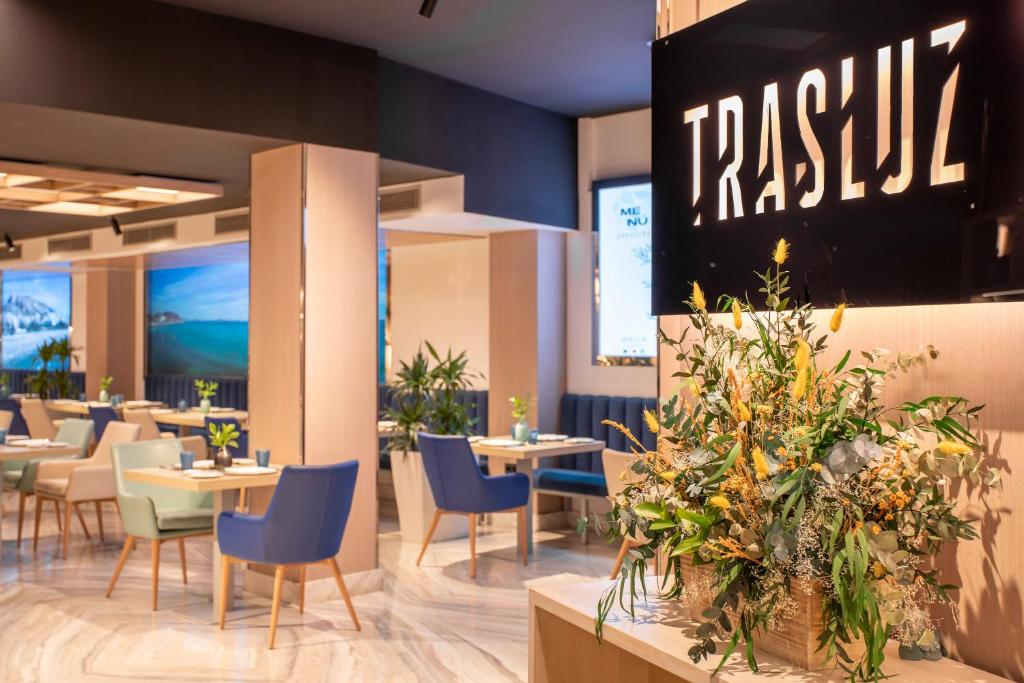 Mūsdienīgs restorāna interjers ar ziedu kompozīciju un zīmolu "TRASLŪZ". Telpā ir komfortabli galdi un krēsli, kā arī lieli ekrāni ar ainavām.