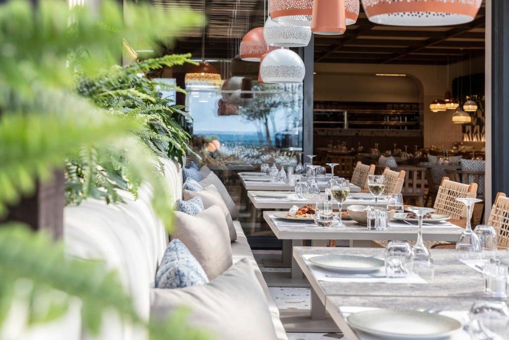 Restorāna interjers ar elegantiem galdiem, modernām lampām un zaļām augu dekorācijām. Skats uz jūru papildina relaksējošo atmosfēru.