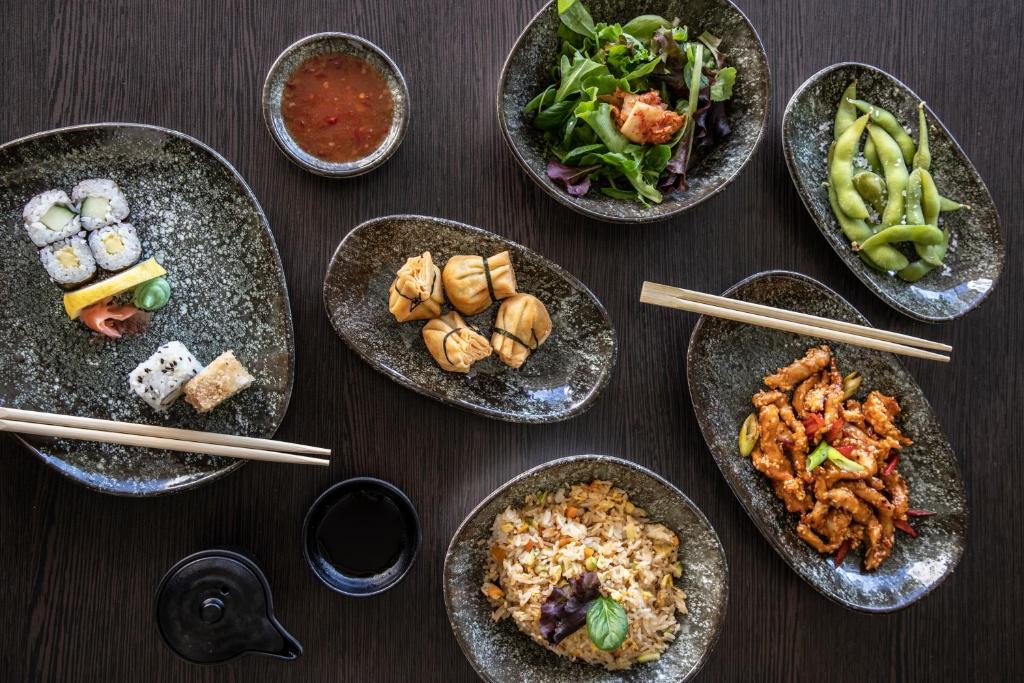Dažādu Āzijas ēdienu kolekcija, tostarp suši, dim sum, salāti, edamame un cepta rīsu porcija, pasniegta uz melnām šķīvēm ar ēdamkarotēm un nūdelēm.