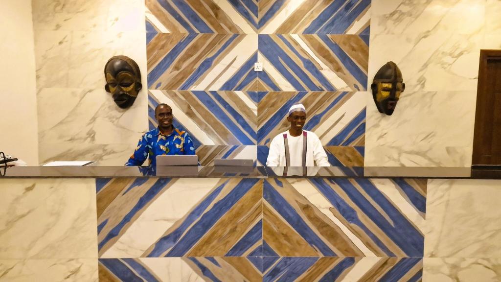 Recepcija ar diviem darbiniekiem, kuri stāv pie moderna dizaina galda ar ģeometriskām sienām un tradicionālām maskām.