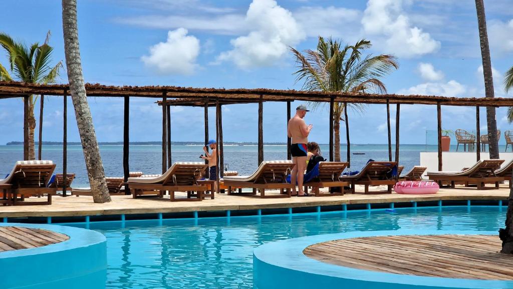 Tropu kūrorts ar baseinu, sauļošanās krēsliem un skatu uz jūru, kur cilvēki atpūšas un fotogrāfē. Ideāla vieta relaksācijai un brīvdienām.