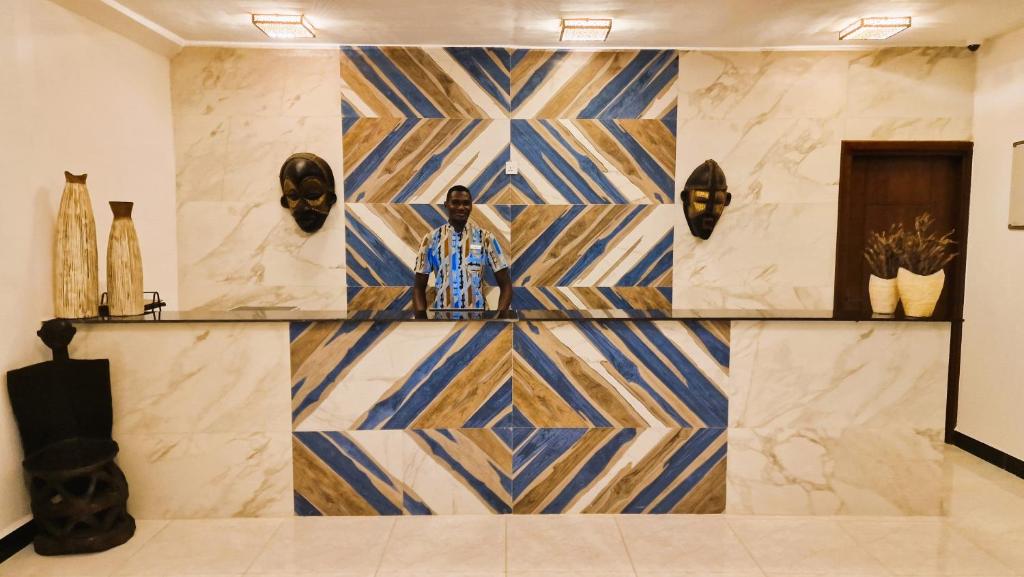 Recepcija ar modernu dizainu, kas ietver ģeometriskas formas un dekoratīvus elementus, piemēram, maskas un stāvošas figūras. Apskatāms viesmīlis, kurš stāv aiz reģistratūras letes.