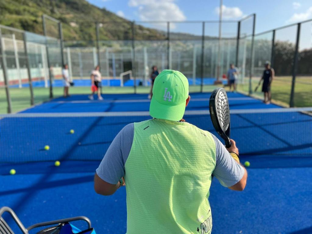 Atsvaidzinoša padela spēles aina, kurā redzams spēlētājs ar zaļu cepuri un sportisku tērpu, gatavojoties izsist bumbu. Fons ir sporta laukumi ar citiem spēlētājiem un saulaina daba.