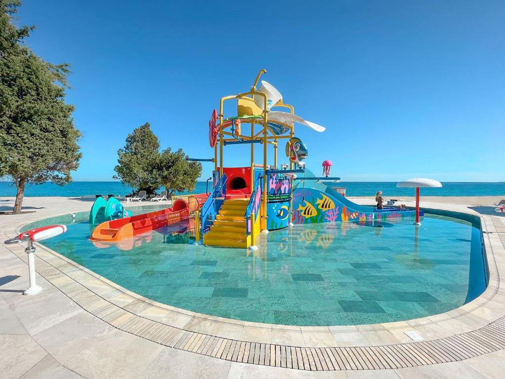 Krāsains bērnu ūdens atrakciju komplekss pie jūras ar slidkalniņiem un rotaļu elementiem, kas atrodas skaidrā ūdenī. Fons veido saulains laiks un zila jūra.