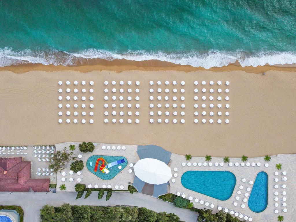 Pludmales skats no augšas ar saulessargiem, kas sakārtoti līnijās, un baseinu tuvumā. Ūdeņi ir dzidri un zili, bet smiltis ir zeltainas.