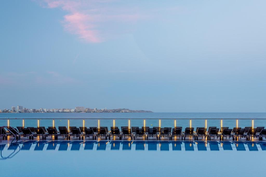 Mūsdienīgs baseins ar sauļošanās krēsliem, kas izstiepti gar ūdens malu, ar skatu uz mierīgu jūru un pilsētu tālumā. Ideāla vieta relaksācijai un atpūtai.
