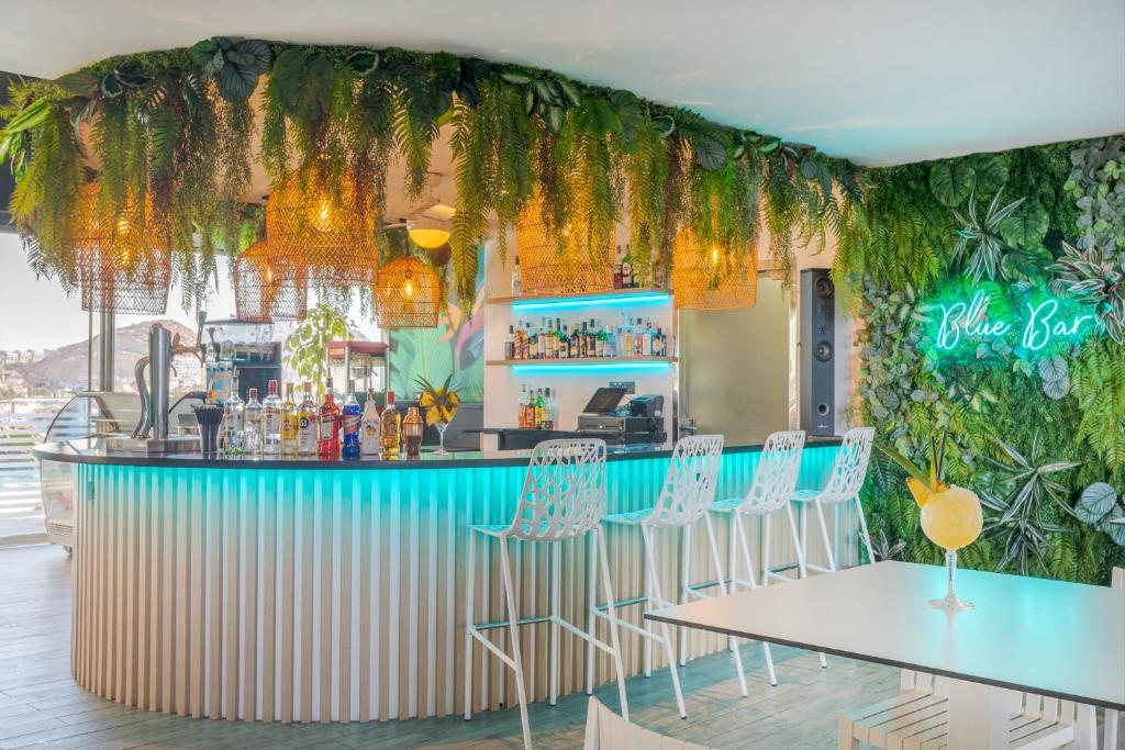 Mūsdienīgs bārs ar zaļumiem un neona uzrakstu "Blue Bar", kurā ir stilīgi krēsli un labi sakārtota dzērienu plaukti. Atmosfēra ir relaksējoša un svaiga.