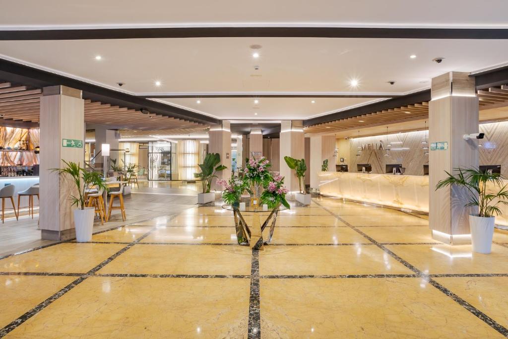 Mūsdienīga viesnīcas recepcija ar elegantu interjeru, gaismas efektu un zaļām augiem. Plaša foajē ar marmora grīdu un koka elementiem, kas rada viesmīlīgu atmosfēru.