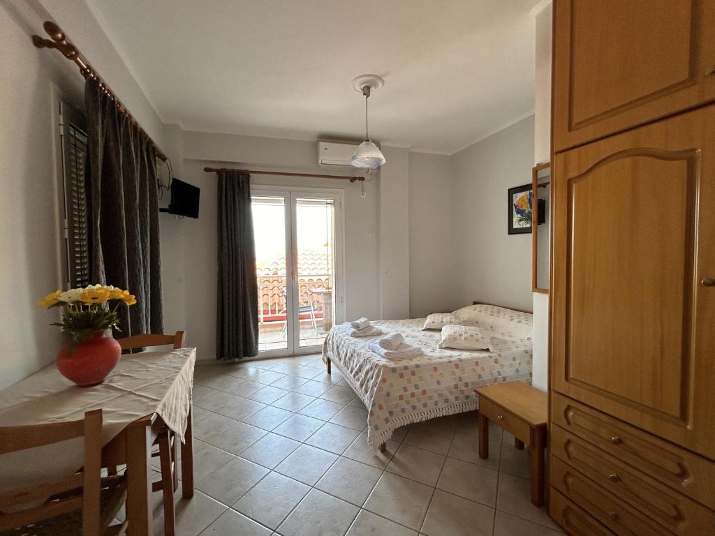 Gaisma un mājīga viesnīcas numuriņa iekārtojums ar divguļamo gultu, koka mēbelēm un balkona durvīm, kas ved uz ārpusi. Uz galda ir ziedu kompozīcija, radot mājīgu atmosfēru.