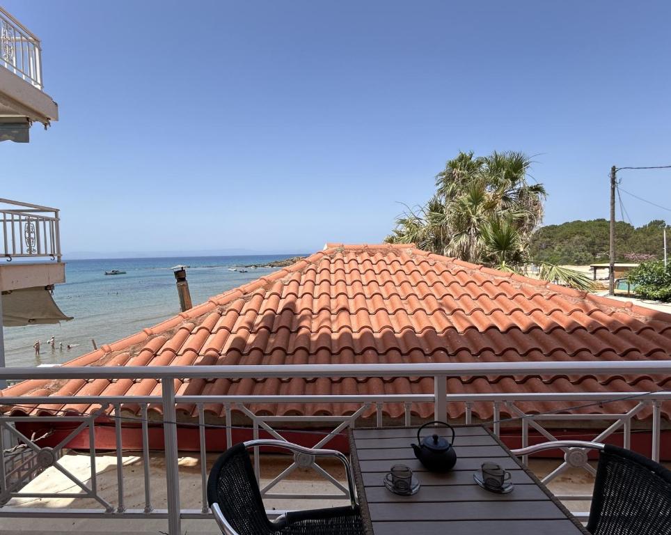 Skats no balkona uz jūru ar terases galdu, kurā redzams tējas komplekts. Fons veido jumts ar sarkanu flīžu segumu un palmas, kas piešķir tropisku atmosfēru.