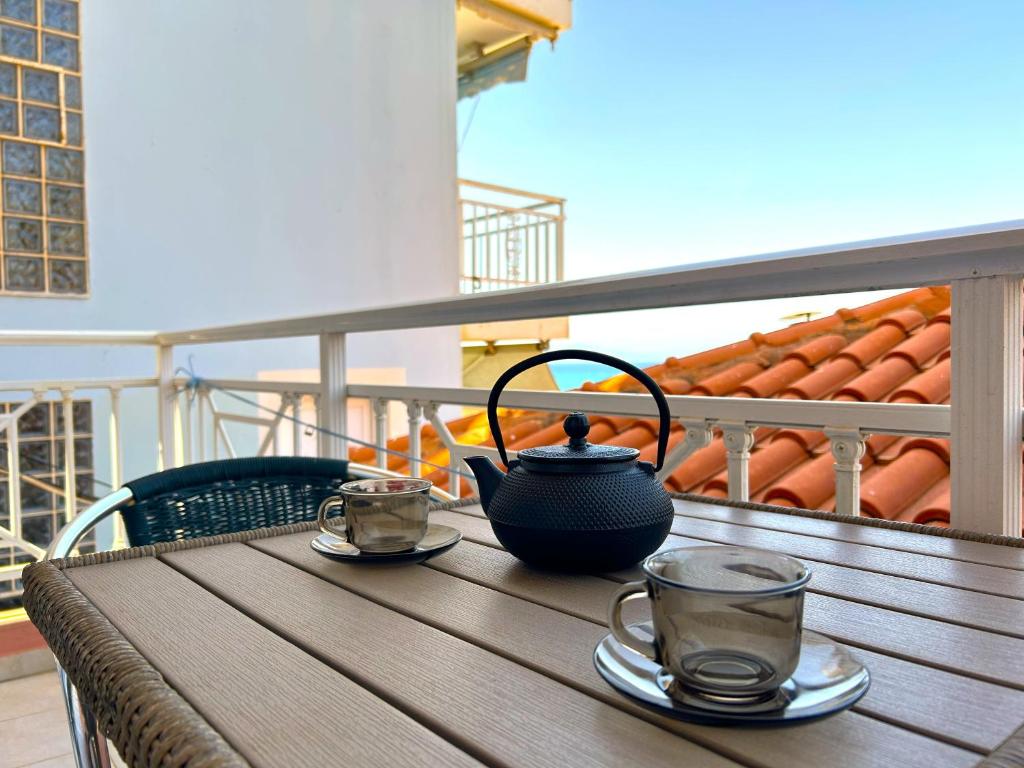 Terase ar tējas komplektu, melnu tējas podu un divām caurspīdīgām krūzēm, fonā redzami sarkanie jumti un skaidra debess.