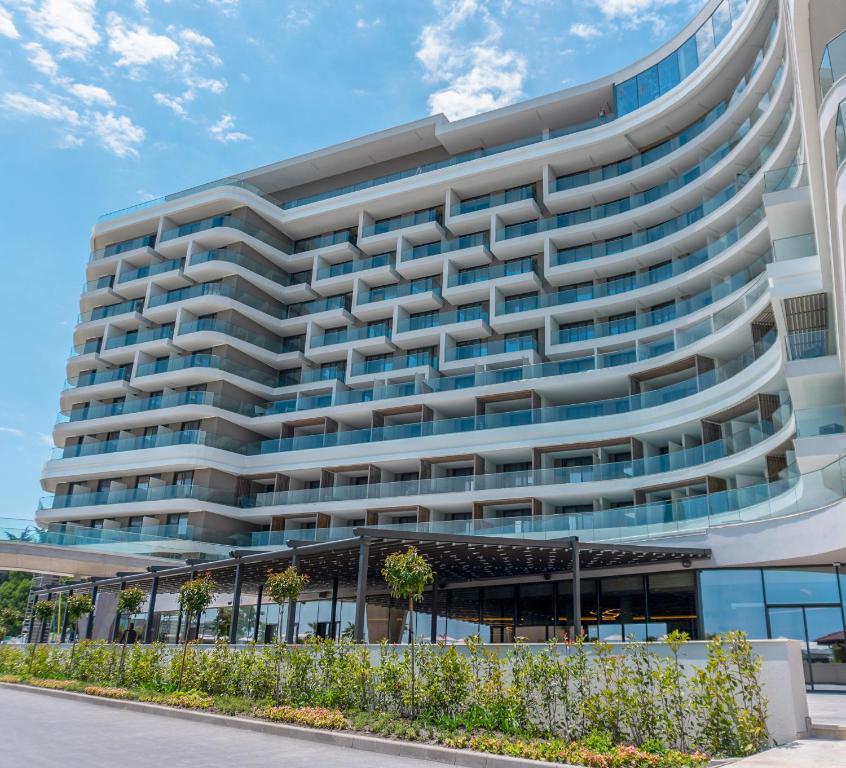 Mūsdienīgs viesnīcas ēkas dizains ar plašiem balkoniem un stikla fasādi, kas atrodas saulainā dienā. Apkārtne ir sakopta ar zālienu un dekoratīvām augiem.