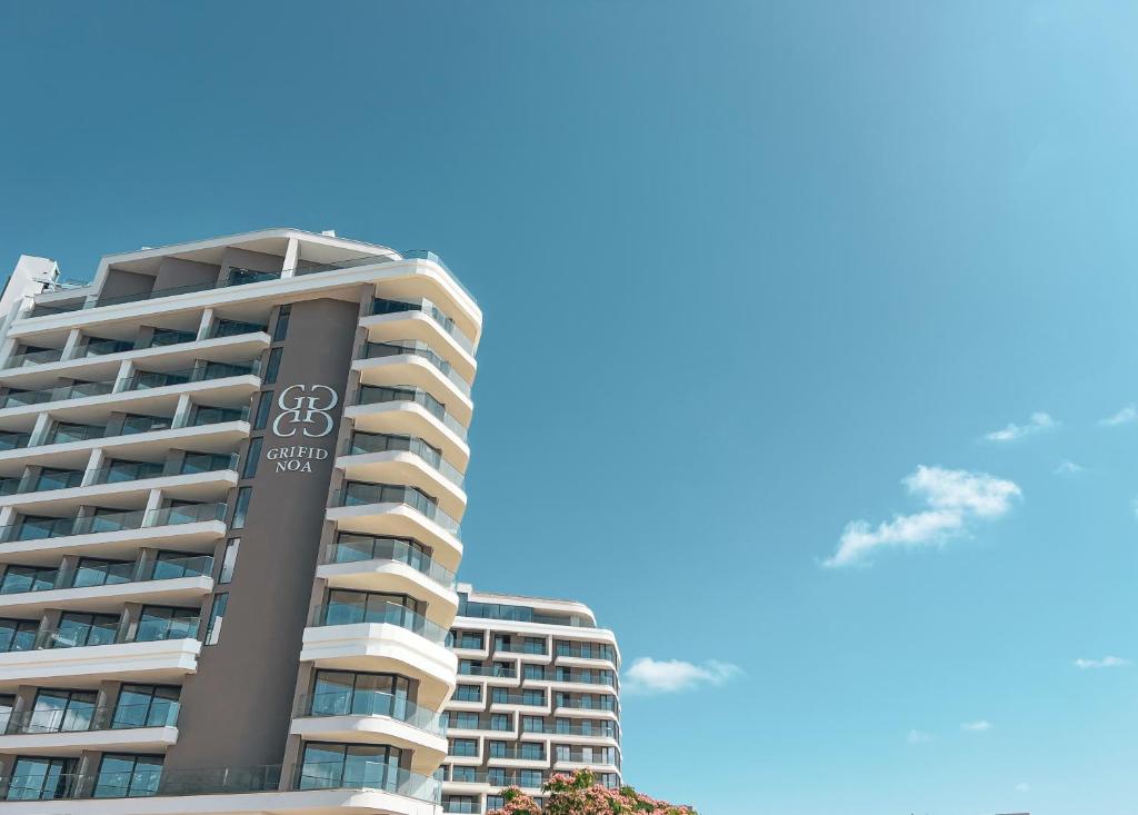 Mūsdienīga viesnīca ar elegantu arhitektūru un logotipu uz fasādes, zem skaidra zila debesu fona.