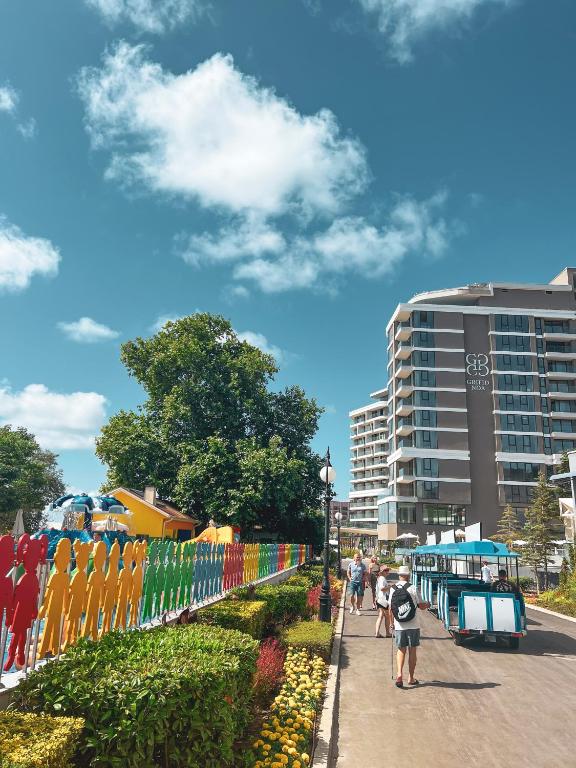 Krāsaina pastaigu zona ar cilvēku figūrām un viesnīcas ēku fonā, saulaina debess un zāles apstādījumi. Ideāla vieta atpūtai un pastaigām.